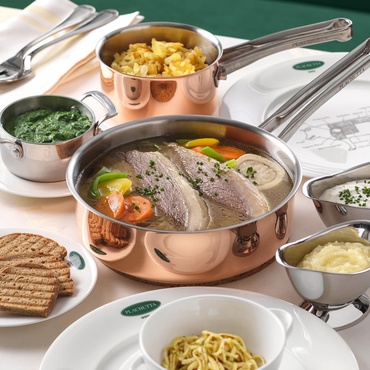 A pot of cooked beef with vegetables, surrounded by side dishes and sauces on a white tablecloth.