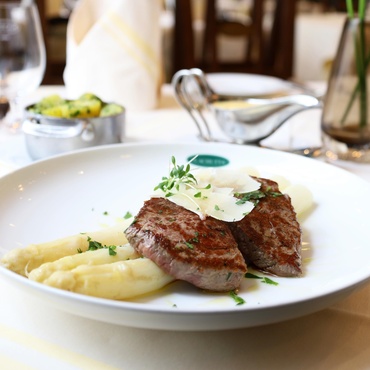 Grilled steak with shaved cheese, white asparagus and mashed potatoes on a white plate in a restaurant setting.